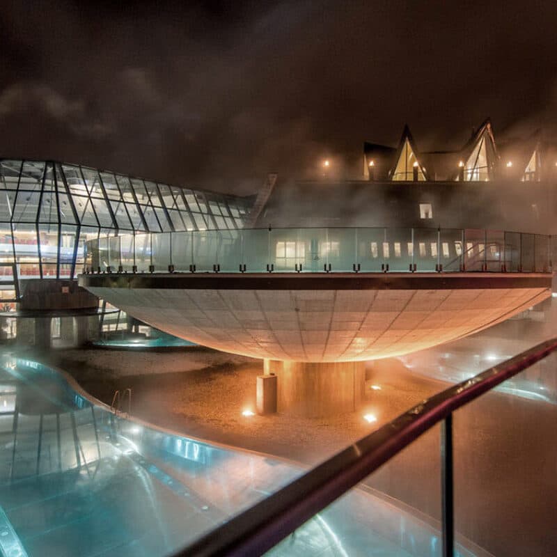 Ein modernes architektonisches Meisterwerk: Die UFO-förmige Therme schwebt anmutig über einem ruhigen Becken, beleuchtet von warmen Lichtern, die durch den nächtlichen Nebel tanzen. Im Hintergrund bilden dreieckige Glasformationen einen markanten Kontrast zu dieser unwirklichen Szenerie.
