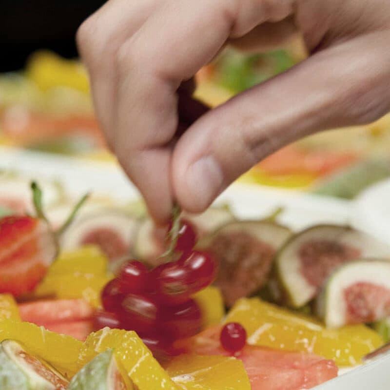 Eine Hand arrangiert kunstvoll rote Beeren auf einer Platte, begleitet von Feigen, leuchtend gelben Früchten und Erdbeeren im Hintergrund, wodurch eine visuelle Therme köstlicher Fülle entsteht.