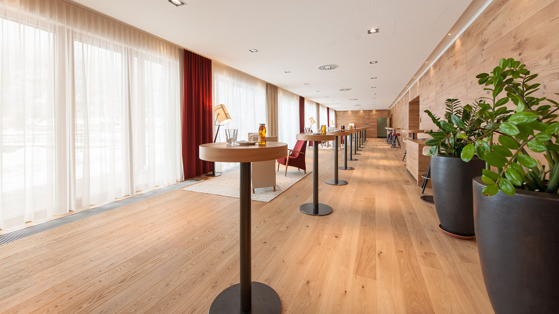 Ein moderner Konferenzraum, ideal für Meetings. Er verfügt über Holzböden, hohe runde Tische, Stühle und große Fenster mit transparenten Vorhängen. Pflanzen und eine Reihe schräger Wandleuchten unterstreichen die Atmosphäre.