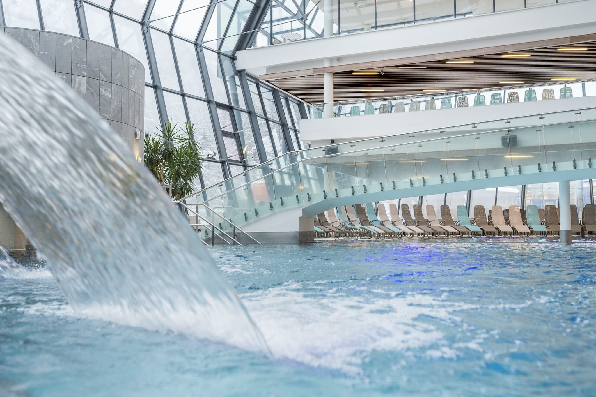 Der Innenpoolbereich, der einer luxuriösen Therme ähnelt, verfügt über eine große Glaskuppel. Ein sanfter Wasserfall stürzt in den Pool, während auf einer oberen Ebene Reihen leerer Liegestühle auf die Gäste warten.