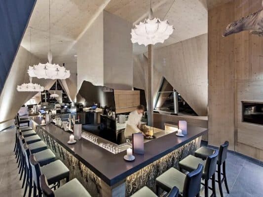 Moderne Bar mit hohen Decken, dreieckigen Fenstern und Holzakzenten. Perfekt für zwanglose Treffen. Hinter der Theke, umgeben von Hockern und Hängelampen, mixt ein Barkeeper Drinks.