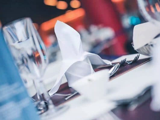 Ein gedeckter Tisch mit einer gefalteten weißen Serviette, Silberbesteck und einem Weinglas, leicht unscharf – fängt perfekt die Eleganz ein, die man oft bei gehobenen Treffen findet.