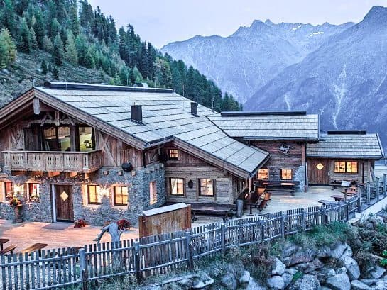 Eine rustikale Holzhütte mit erleuchteten Fenstern liegt auf einem Hügel, umgeben von Bergen und mit einem Zaun davor, und bietet den perfekten Rückzugsort für vertrauliche Treffen.