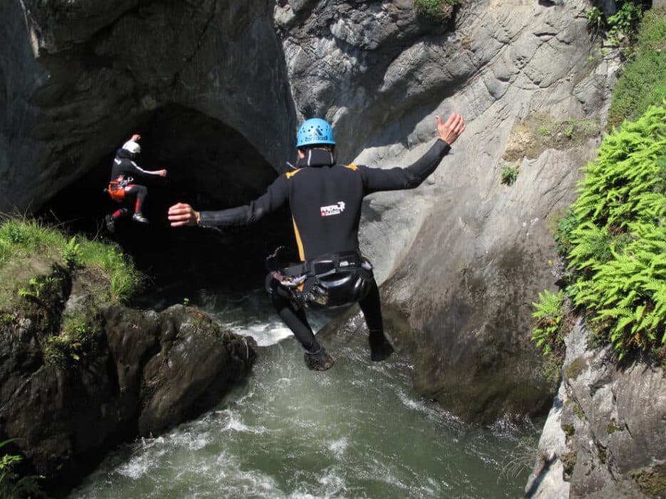Zwei Menschen in Neoprenanzügen und Helmen springen in eine enge Felsschlucht, wo das darunter fließende Wasser eine natürliche Kulisse für unerwartete Begegnungen mit Abenteuern schafft.