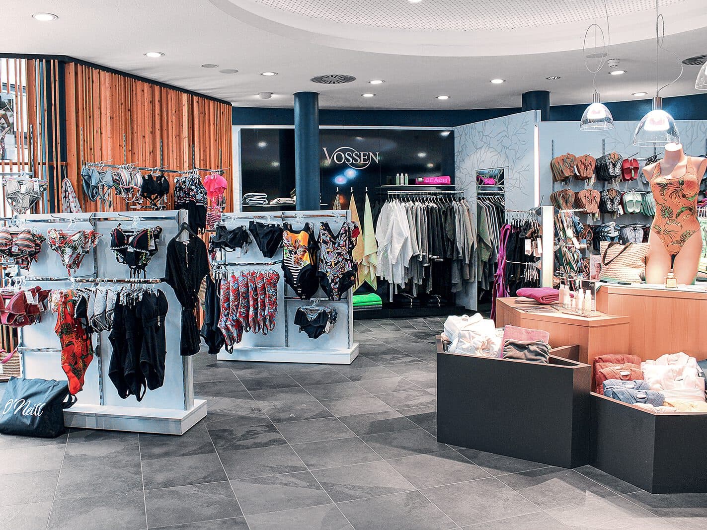 Der Laden besticht durch farbenfrohe Bademoden-Auslagen und schicke Kleiderständer, an denen eine stylische Schaufensterpuppe einen Badeanzug präsentiert. Das Thermen-inspirierte Ambiente wird durch den eleganten grauen Fliesenboden und die stimmungsvolle Deckenbeleuchtung noch verstärkt.
