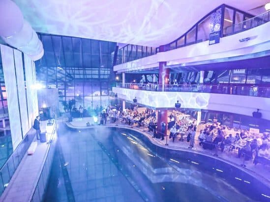 Bei der Indoor-Veranstaltung am Pool genießen die Gäste eine ruhige Atmosphäre, die durch blaues Licht, das Reflexionen an Wänden und Decken wirft, noch verstärkt wird. Mehrere Stockwerke sind sichtbar und schaffen so den idealen Rahmen für vertrauliche Treffen.