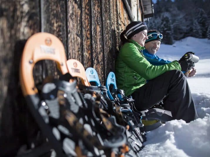 Zwei Personen in Winterkleidung sitzen an einer Holzwand im Schnee, umgeben von mehreren Paar Schneeschuhen. Es scheint, als hätten sie ihr frostiges Treffen unterbrochen, um inmitten der winterlichen Stille ihre Ausrüstung zurechtzurücken oder festzuziehen.