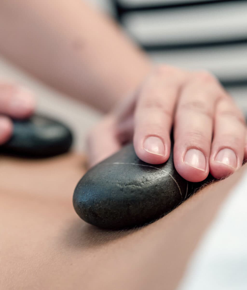 Nahaufnahme einer Person, die eine Hot-Stone-Massage erhält. Zwei Hände legen dunkle Steine auf den Rücken der Person, vor einem unscharfen Spa-Hintergrund.