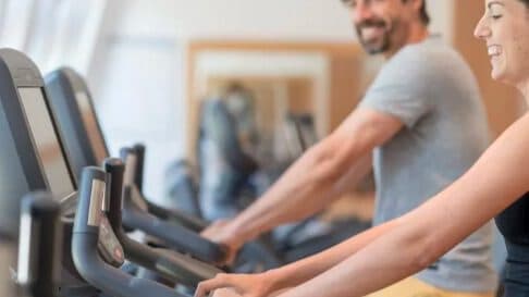 Zwei Personen trainieren in einem Fitnessstudio auf Heimtrainern, lächeln und freuen sich.