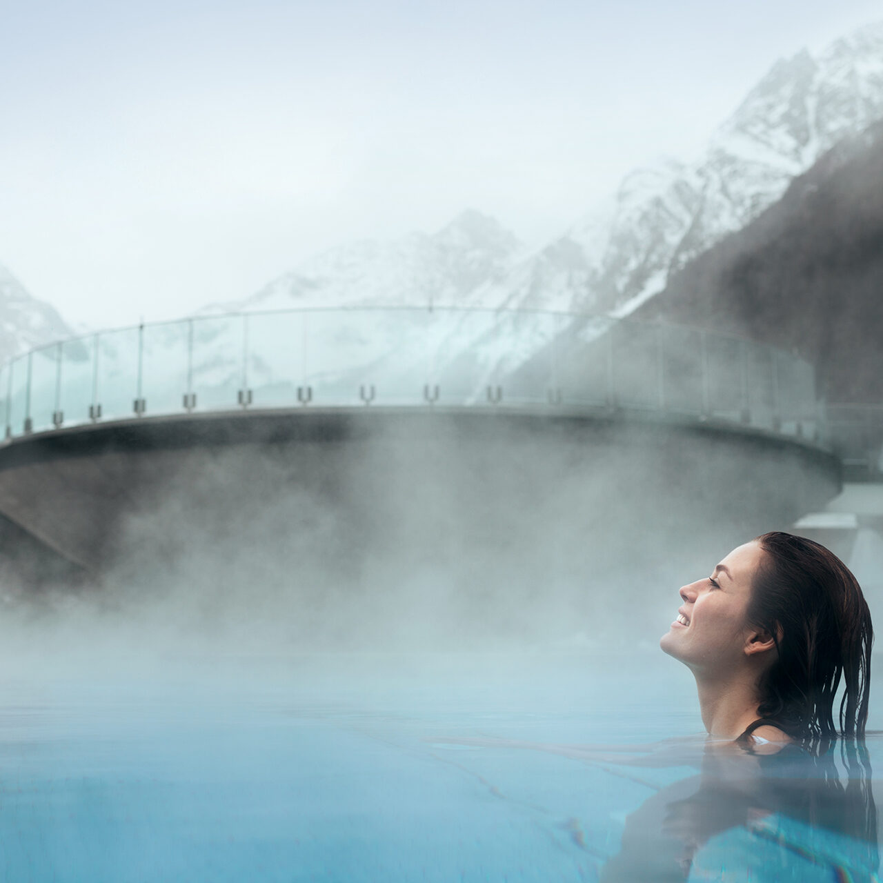 Eine Person entspannt sich in einem dampfenden Außenpool mit schneebedeckten Bergen und einer Glasbrücke im Hintergrund.