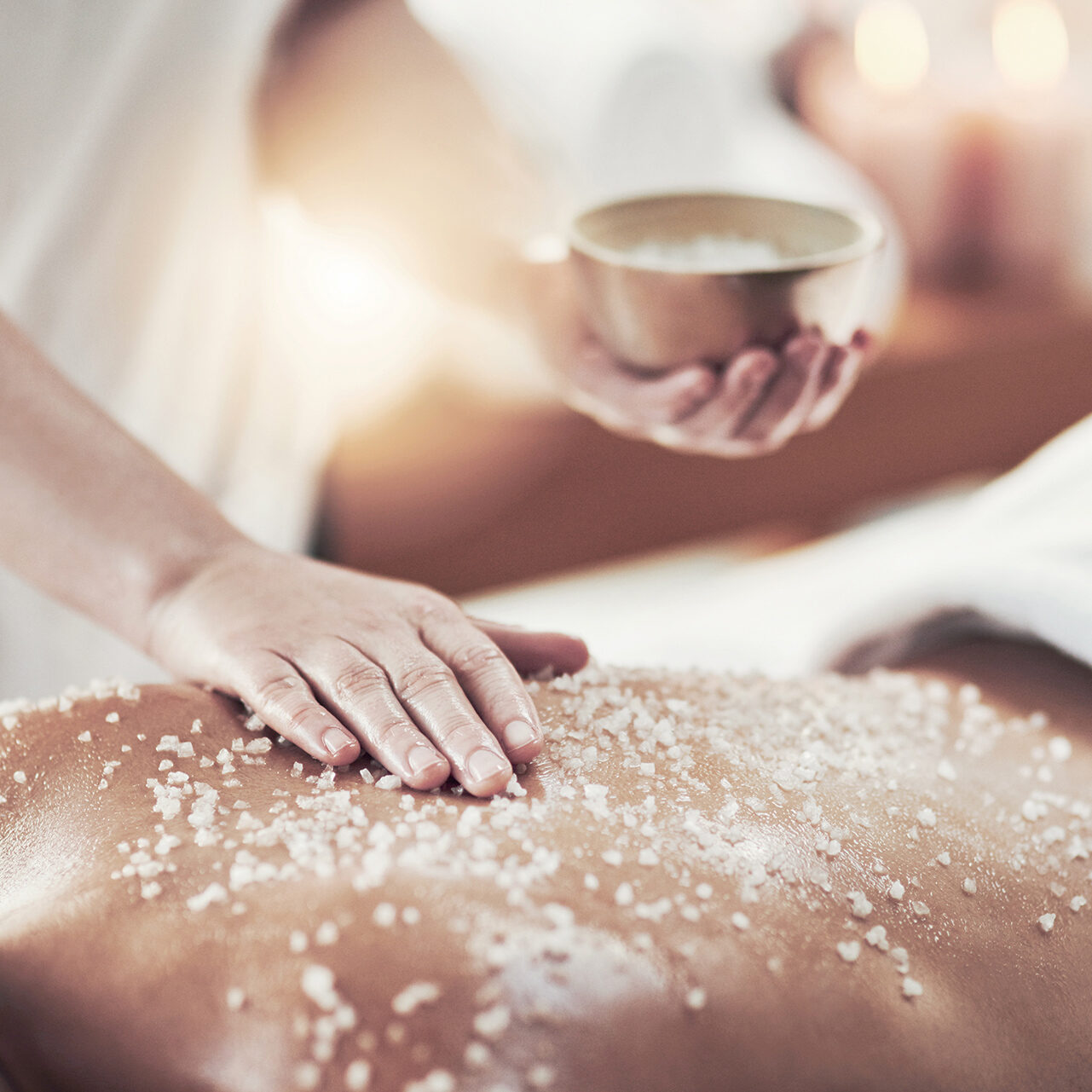 In einem Spa trägt die Hand einer Person Peelingsalz auf den Rücken einer anderen Person auf. In der Nähe steht eine kleine Schüssel, im Hintergrund ist sanftes Kerzenlicht zu sehen.