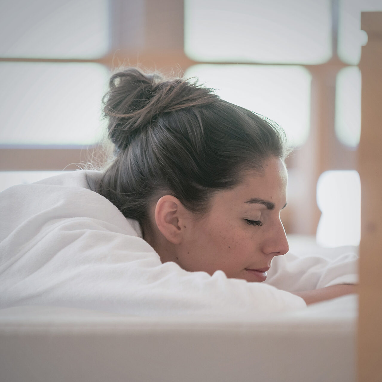 Frau mit dunklem Haar zu einem Dutt und weißem Bademantel liegt mit geschlossenen Augen auf dem Bauch in einem hellen Raum.