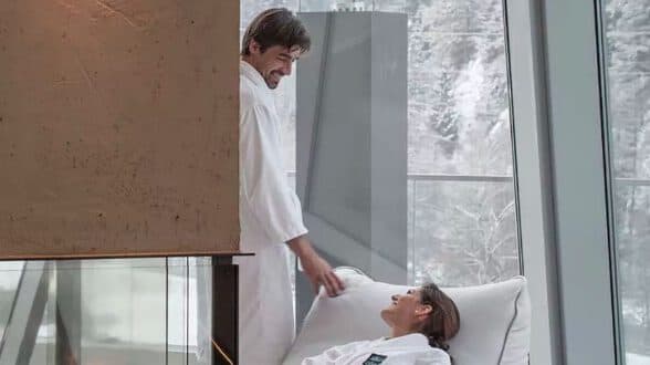 Zwei Menschen in weißen Gewändern entspannen sich an einem großen Fenster mit Blick auf die verschneite Landschaft. Einer steht da und lächelt den anderen an, der auf einem Stuhl liegt.