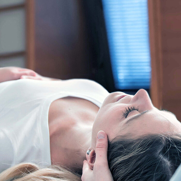 Eine Person liegt mit geschlossenen Augen auf dem Rücken und erhält eine Kopfmassage. Sie trägt ein weißes Hemd. Im Hintergrund sind ein Fenster mit Jalousien und Holzpaneelen zu sehen.