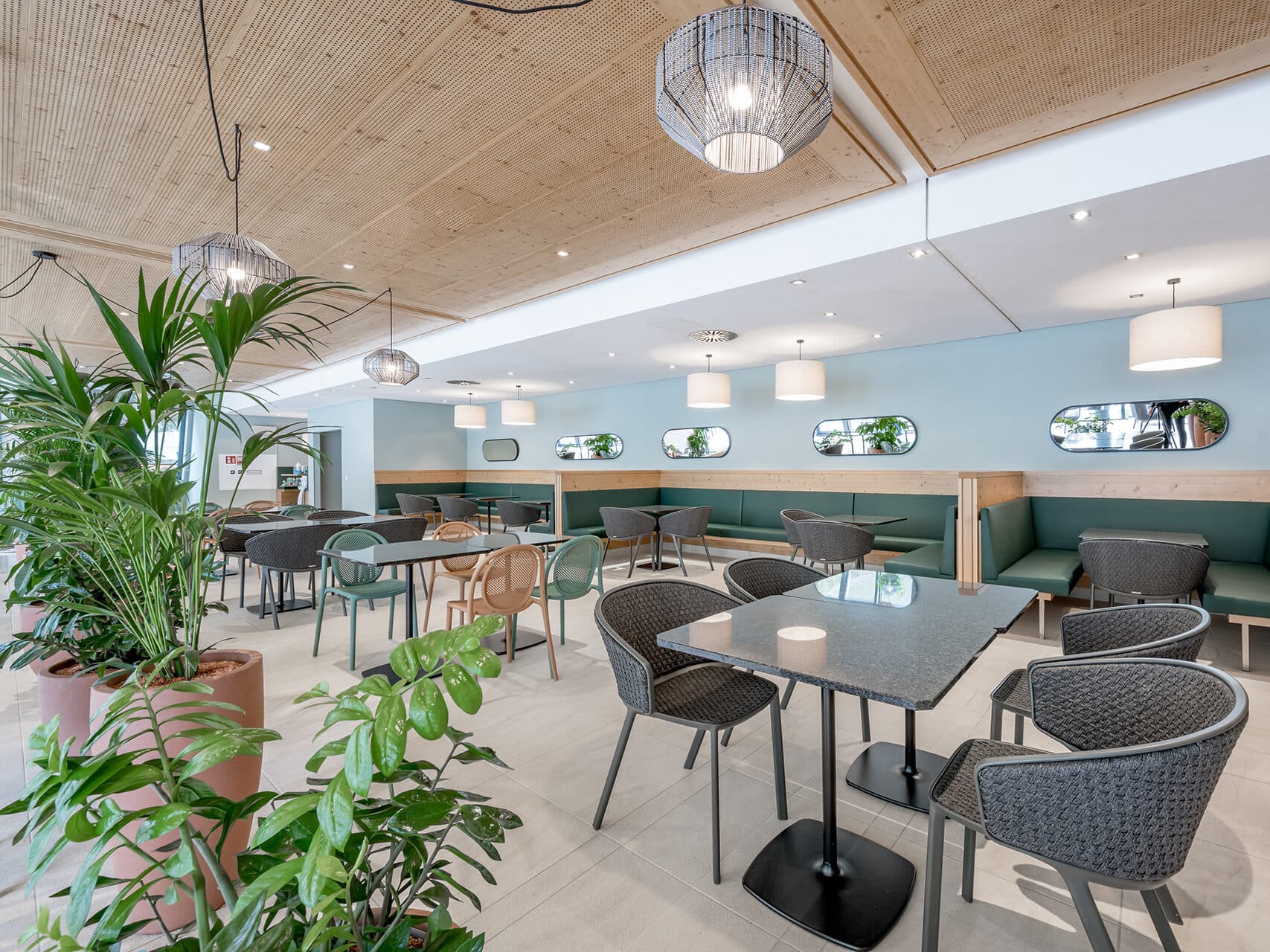 Das moderne Café-Interieur mit grauen Tischen, Korbstühlen und Pendelleuchten schafft einen einladenden Raum für Meetings. Grüne Pflanzen ergänzen die hellblauen und hölzernen Akzente, während Spiegel an der Wand für Tiefe sorgen.