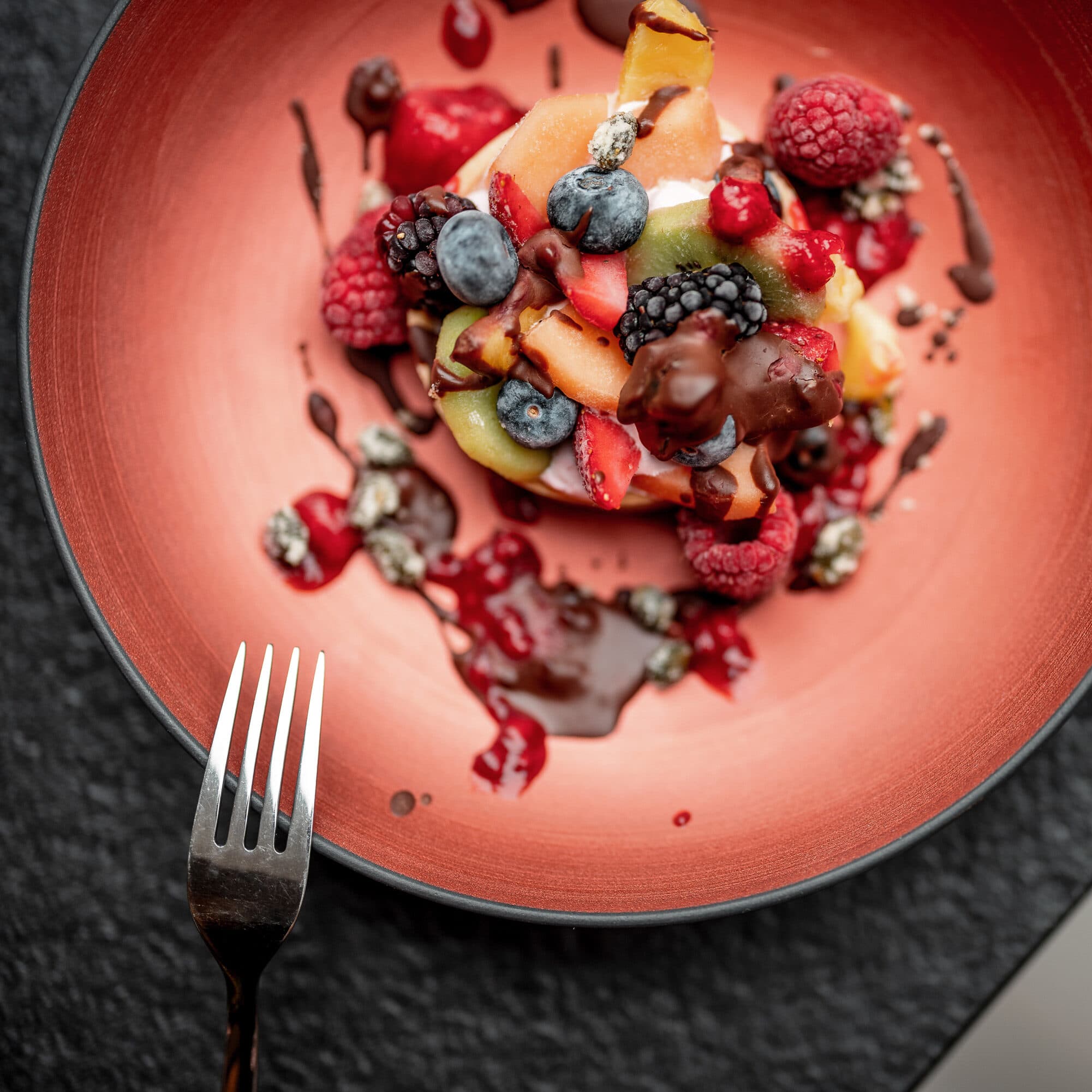 Ein Teller mit verschiedenen frischen Früchten, darunter Beeren und Kiwi, beträufelt mit Schokoladensoße und einer Gabel an der Seite.