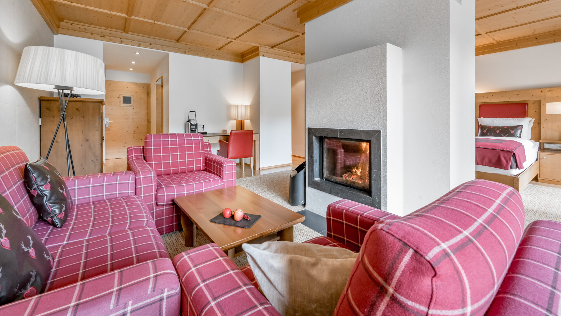 Wohnbereich mit rotkarierten Sesseln, einem Couchtisch mit Äpfeln, einem Kamin und einem sichtbaren Schlafzimmer in einer gemütlichen, modernen Suite mit Holzakzenten.