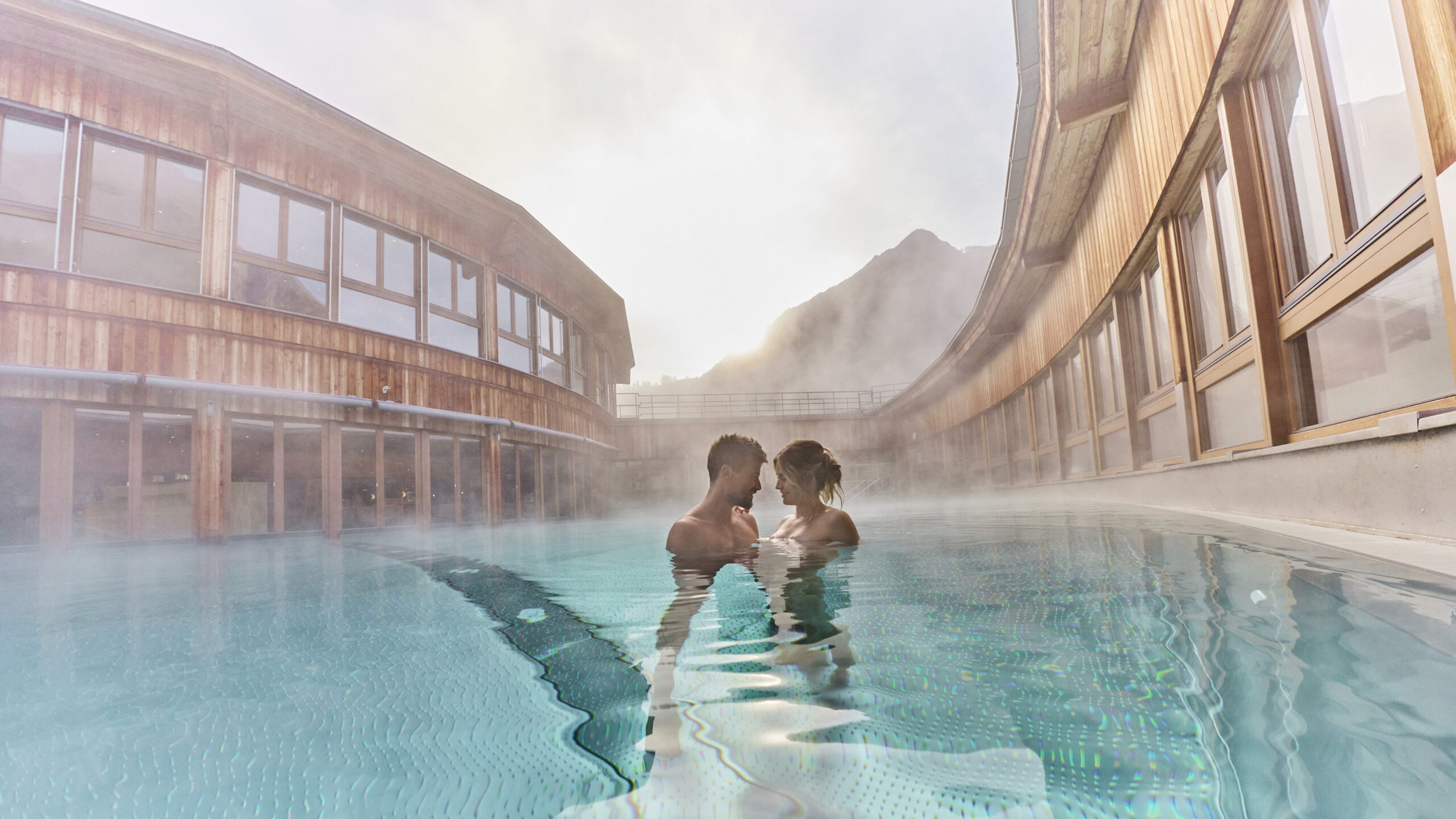 Zwei Personen stehen dicht beieinander in einem dampfenden Freibad, umgeben von modernen Holzgebäuden, mit einem Berg im Hintergrund.