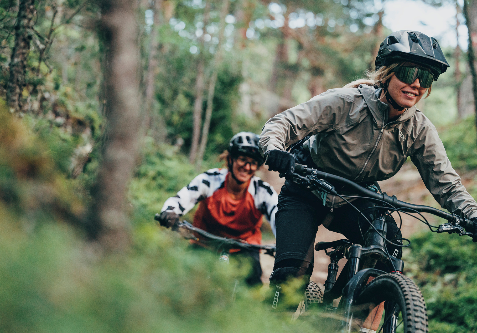 Zwei Personen mit Helmen und Jacken fahren mit dem Mountainbike auf einem Waldweg, wobei eine Person vorausfährt und die andere dicht dahinter folgt.