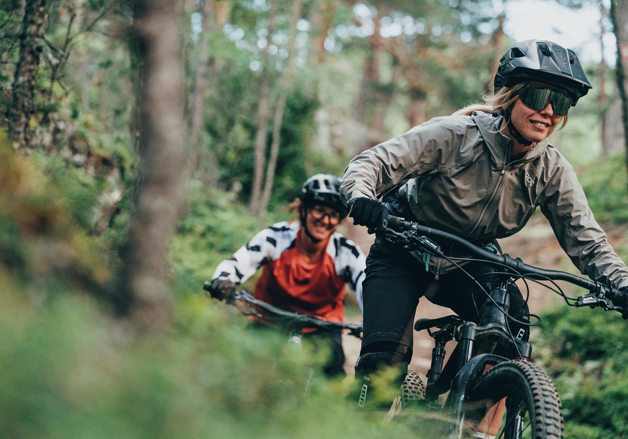 Two people wearing helmets and sunglasses ride mountain bikes on a forest trail, surrounded by trees and greenery.