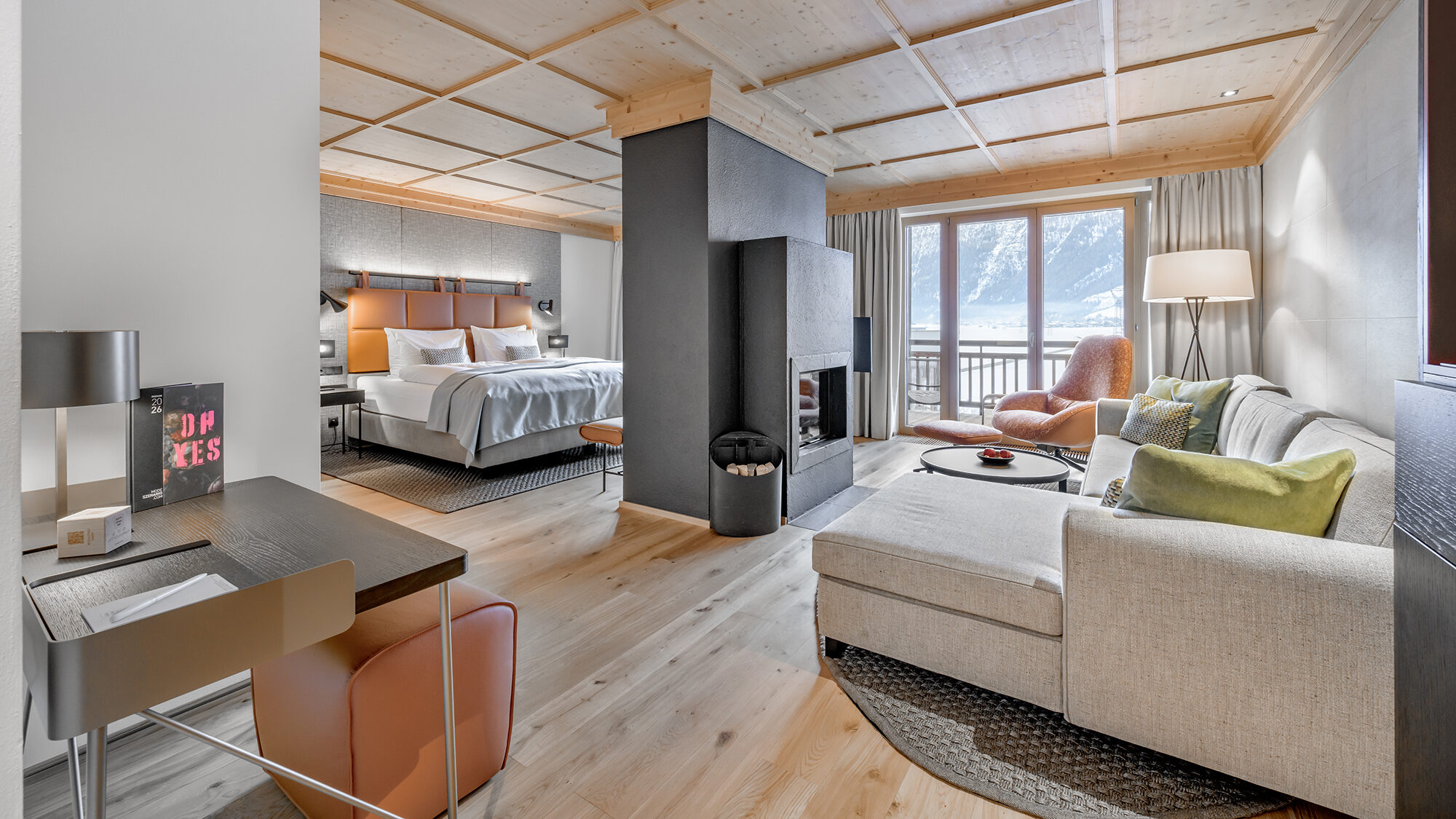 Moderne Hotelsuite mit Kingsize-Bett, Schreibtisch, Sofa, Kamin, Sitzecke und großem Fenster mit Blick auf die Berge.