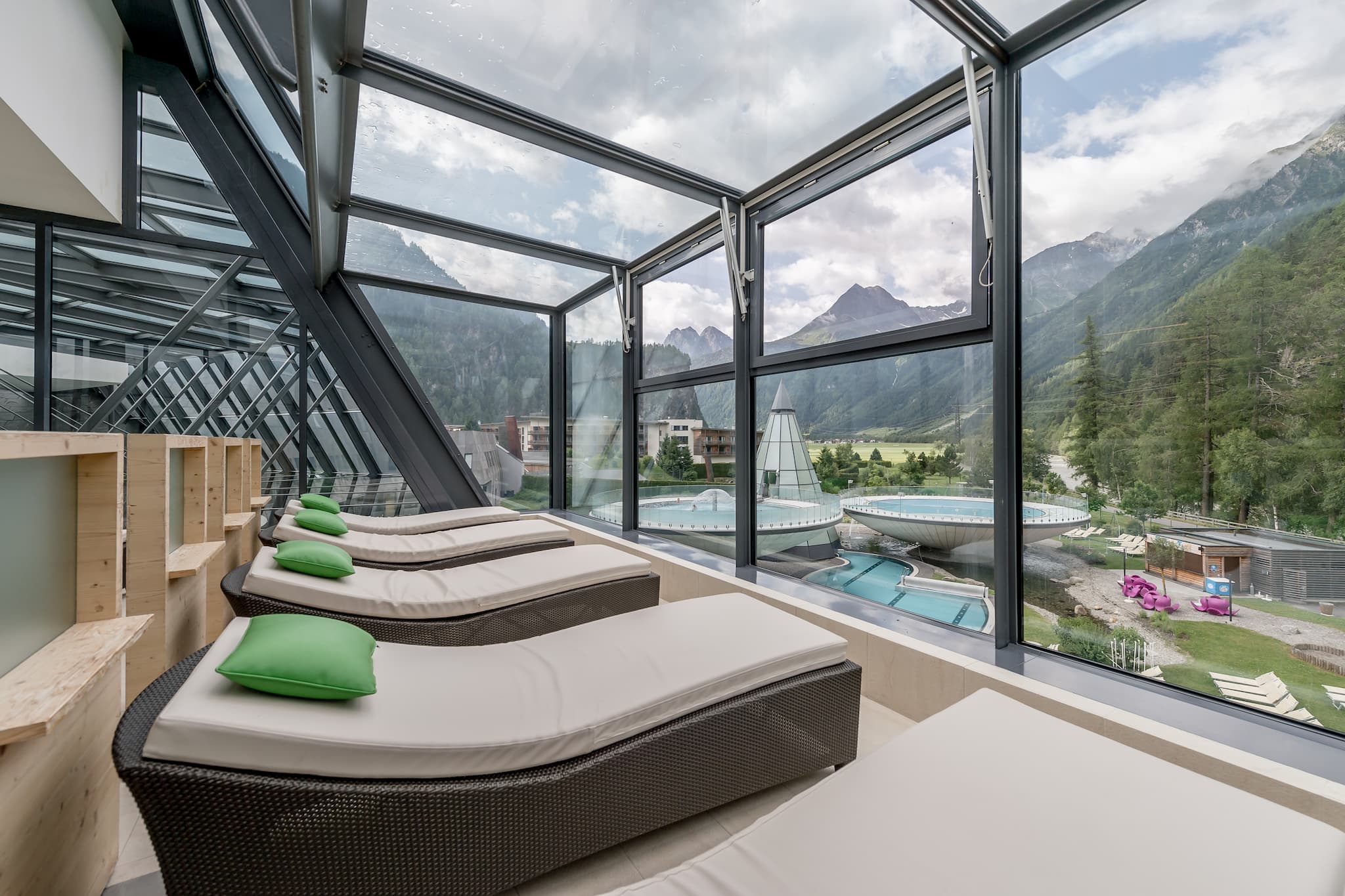 Lounge chairs with green pillows in a glass-enclosed area offer a view of mountains and outdoor pools on a partly cloudy day.