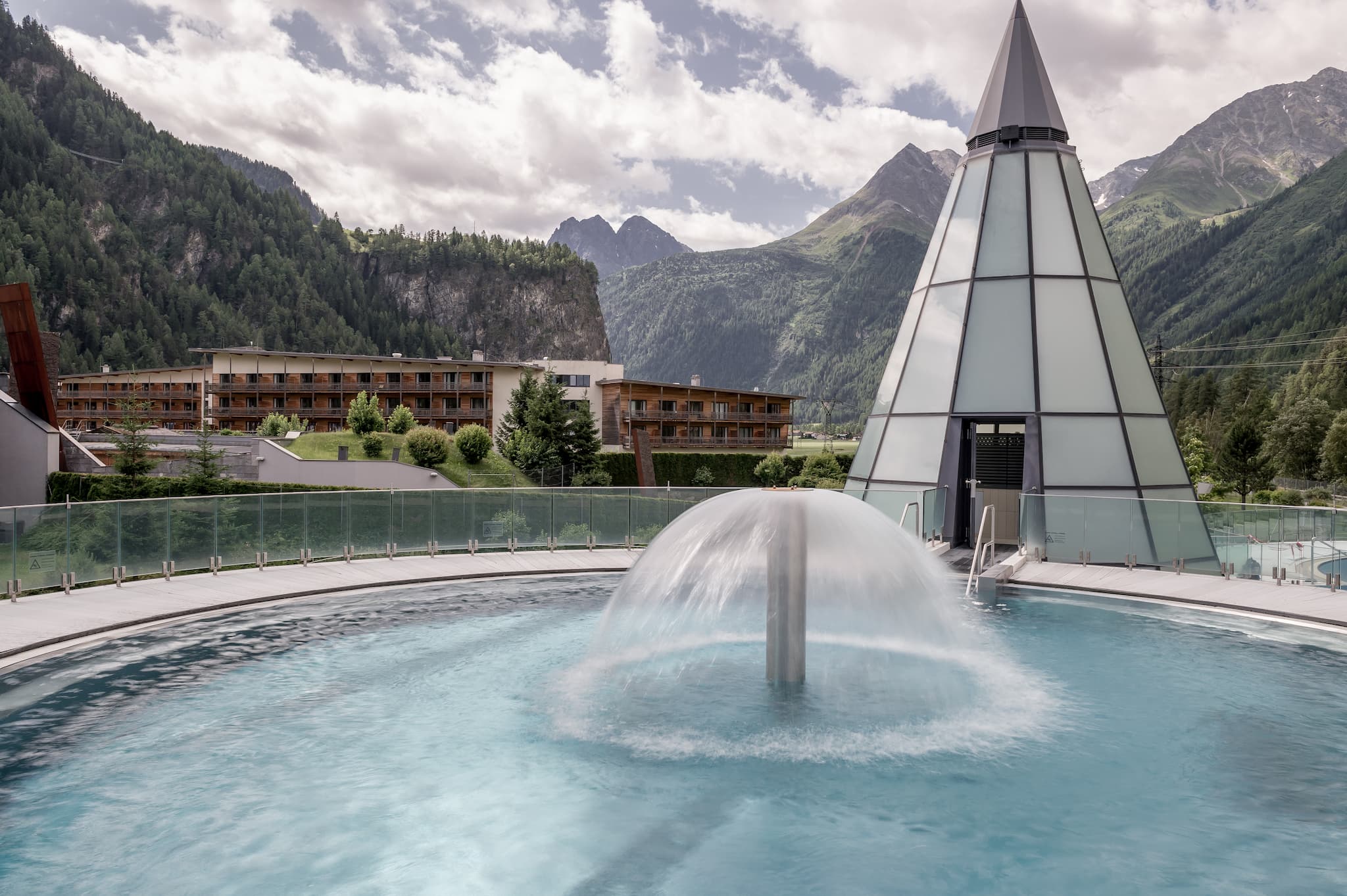 A circular outdoor pool with a central fountain, surrounded by mountains. A glass pyramid structure stands nearby, with a view of buildings in the background.