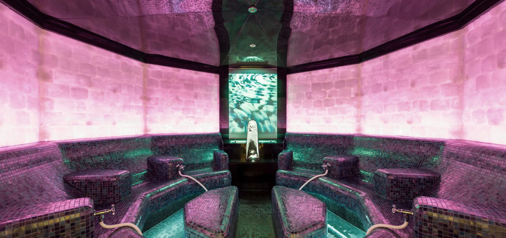 Purple and green themed steam room with mosaic tiled benches, central water fountain, and ambient lighting.