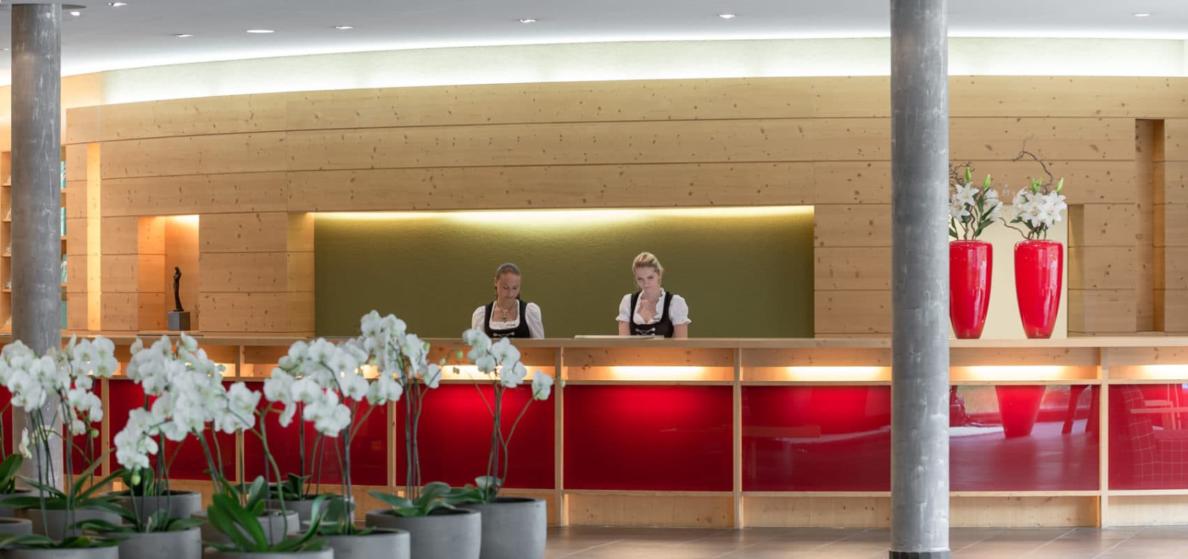 Two employees in uniform stand behind a wooden and red reception desk with orchids in the foreground and red vases on the right.