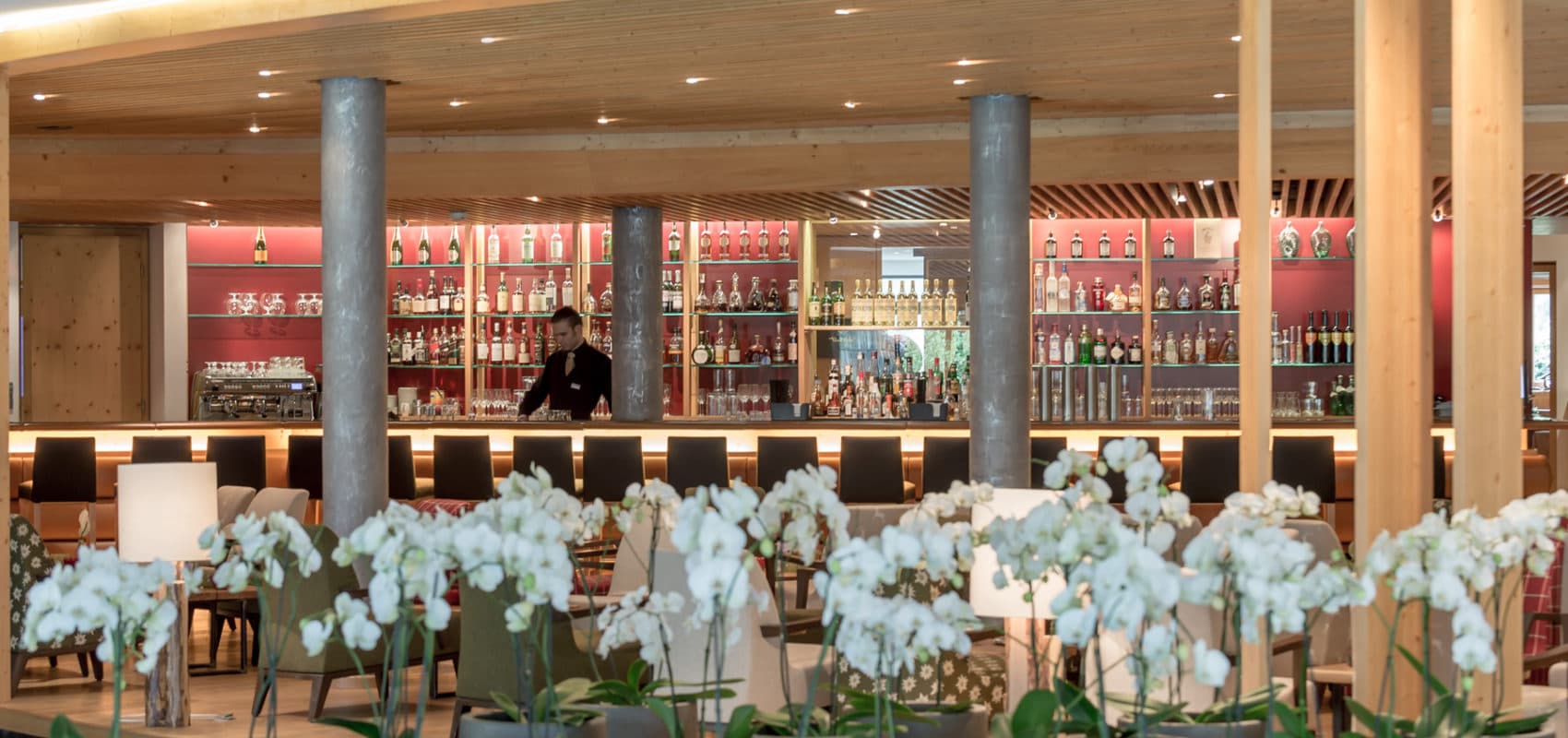 A modern hotel bar with a well-stocked shelf of bottles and glasses, a bartender at the counter, and white orchids in the foreground.