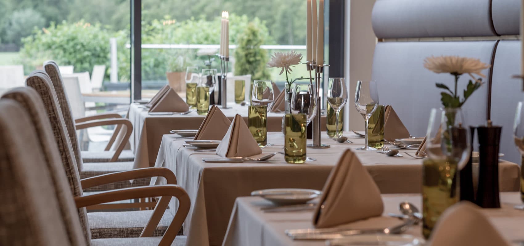 Elegant restaurant setup with tables covered in white tablecloths, neatly arranged tableware, folded napkins, wine glasses, and tall candles. Greenery visible through large windows.