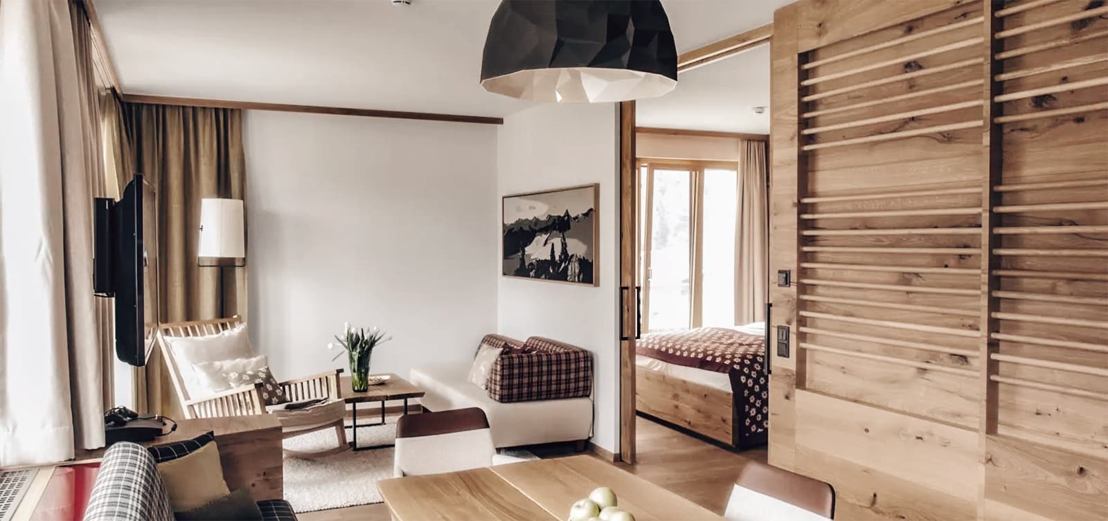 Уютная гостиная с современным креслом, телевизором и искусством на стене. Деревянная раздвижная дверь открывает спальню. Естественный свет освещает пространство.