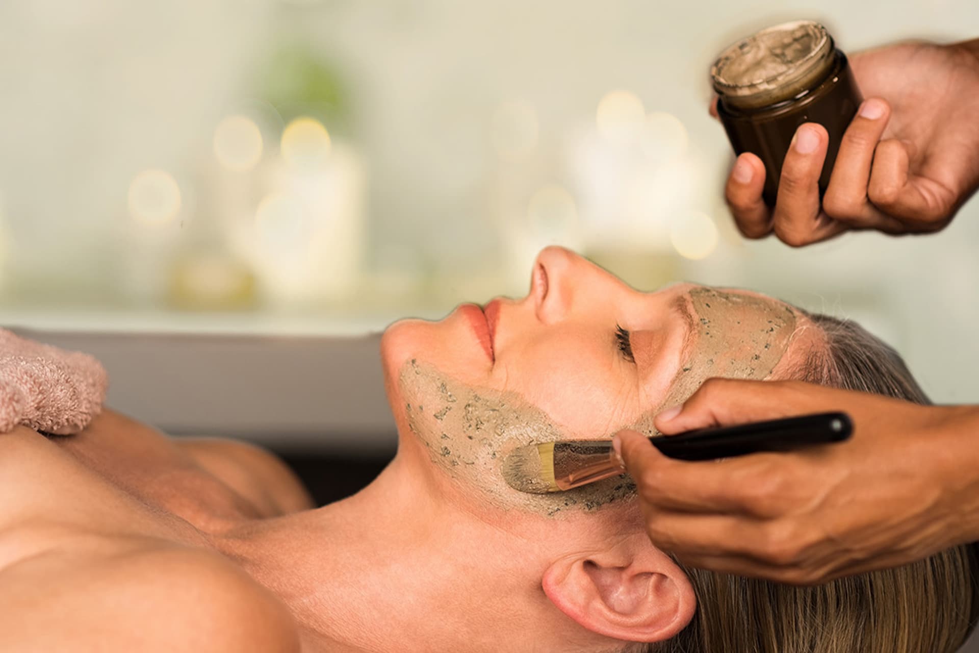 A person relaxes at a thermal spa, enjoying a soothing facial treatment as a brush gently applies a green mask from a small jar.