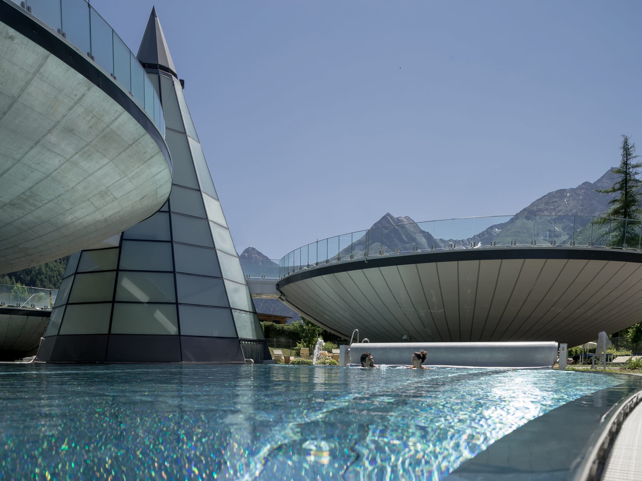 Futuristisches Spa mit pyramiden- und scheibenförmigen Strukturen an einem makellosen Thermenbecken. Im Hintergrund erheben sich majestätische Berge unter einem klaren blauen Himmel.