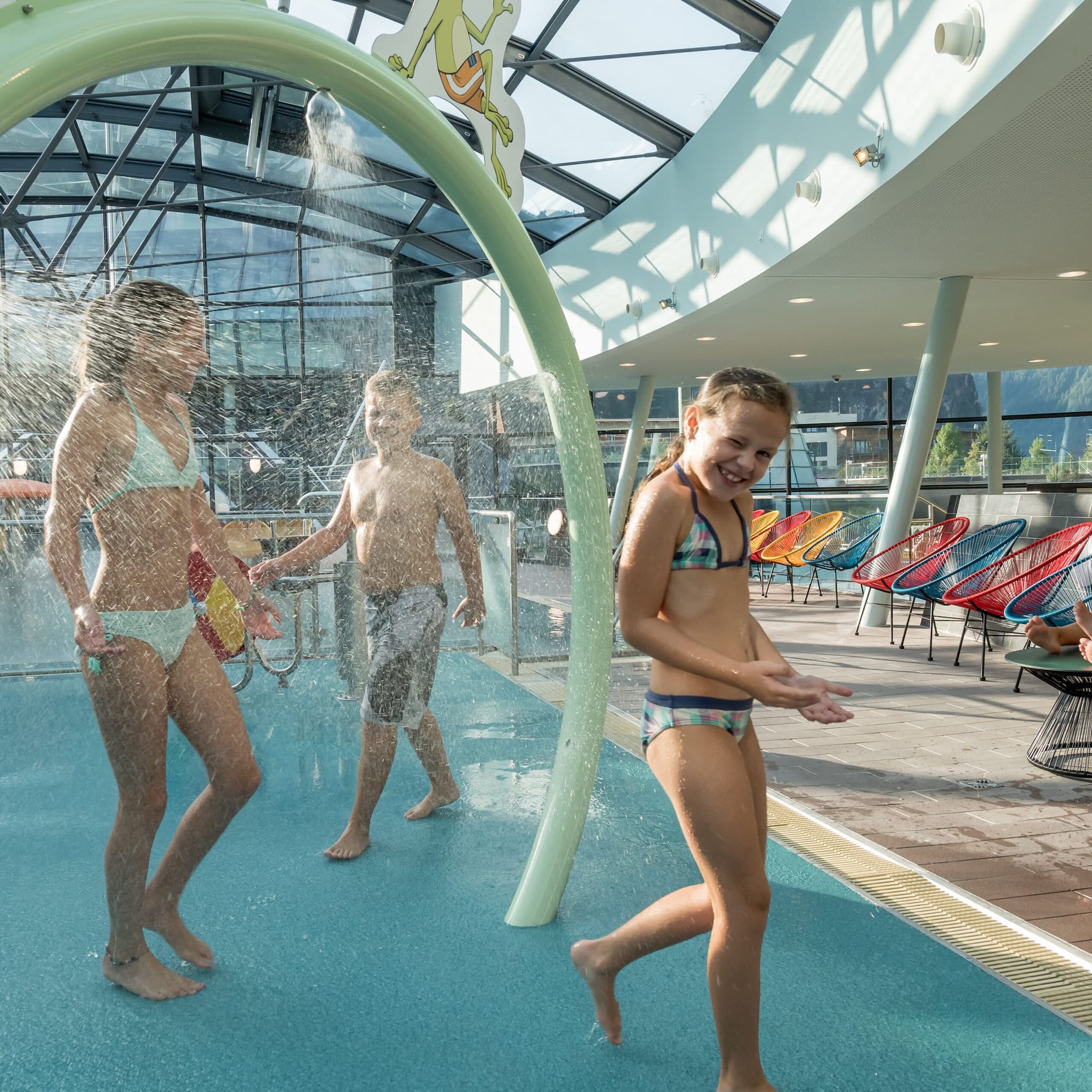 Children gleefully play under a water spray feature in the hotels indoor pool area, with colorful chairs adding to the vibrant backdrop.