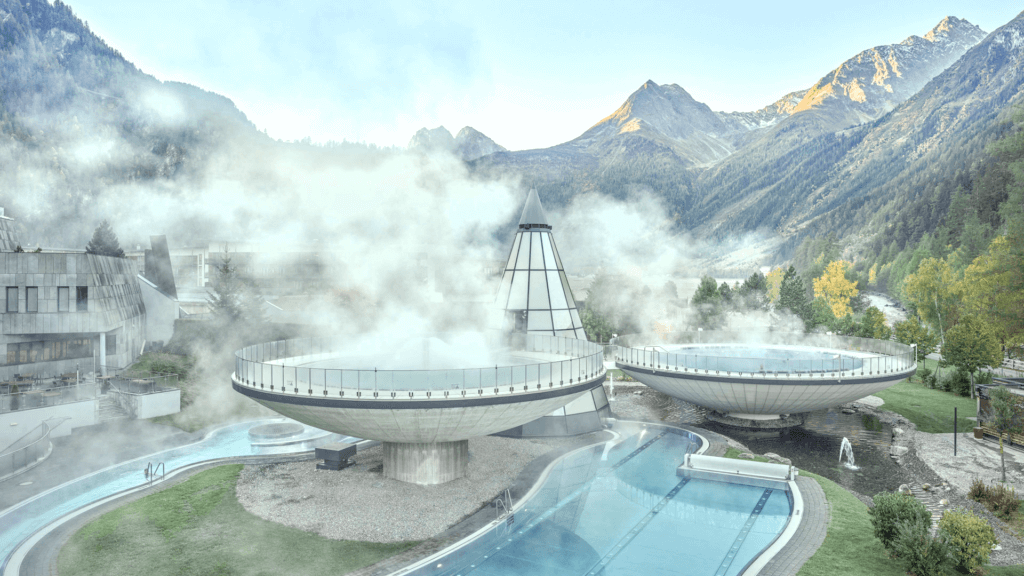 Modern spa complex with steaming circular pools in a mountainous landscape. Surrounding trees and peaks are visible in the background under a clear sky.