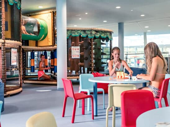 Kinder spielen an Tischen in einem farbenfrohen Indoor-Spielbereich mit Spielstrukturen und Klettergerüsten im Hintergrund.