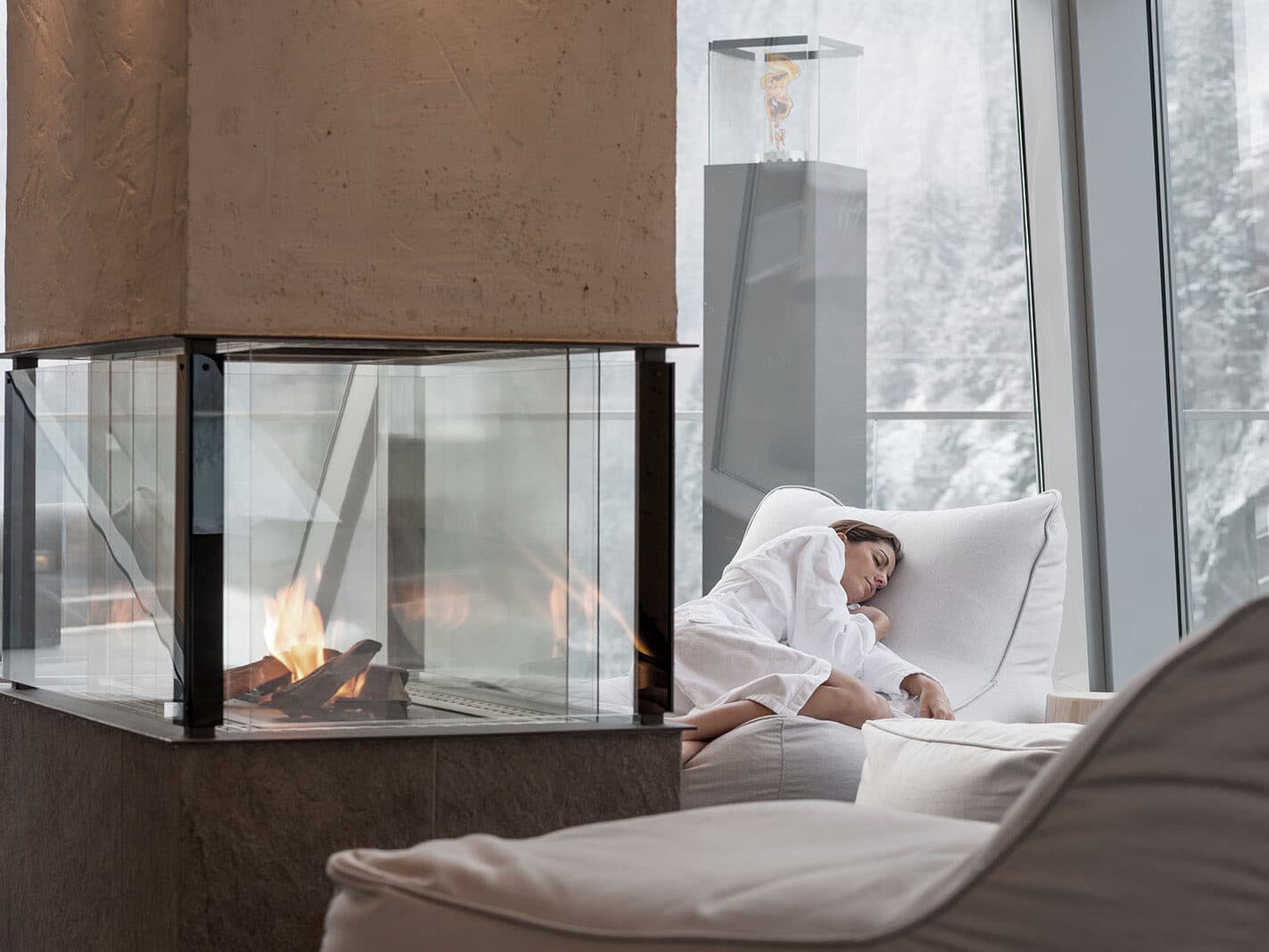 A person in a white robe drifts into slumber in a comfortable chair beside a sleek glass fireplace, enjoying the serenity of their hotel room. Through large windows, they gaze upon the tranquil snowy landscape outside.
