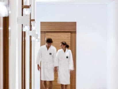 Two people in white robes stroll down a bright hallway with wooden doors, evoking the serene ambiance of a luxurious spa.