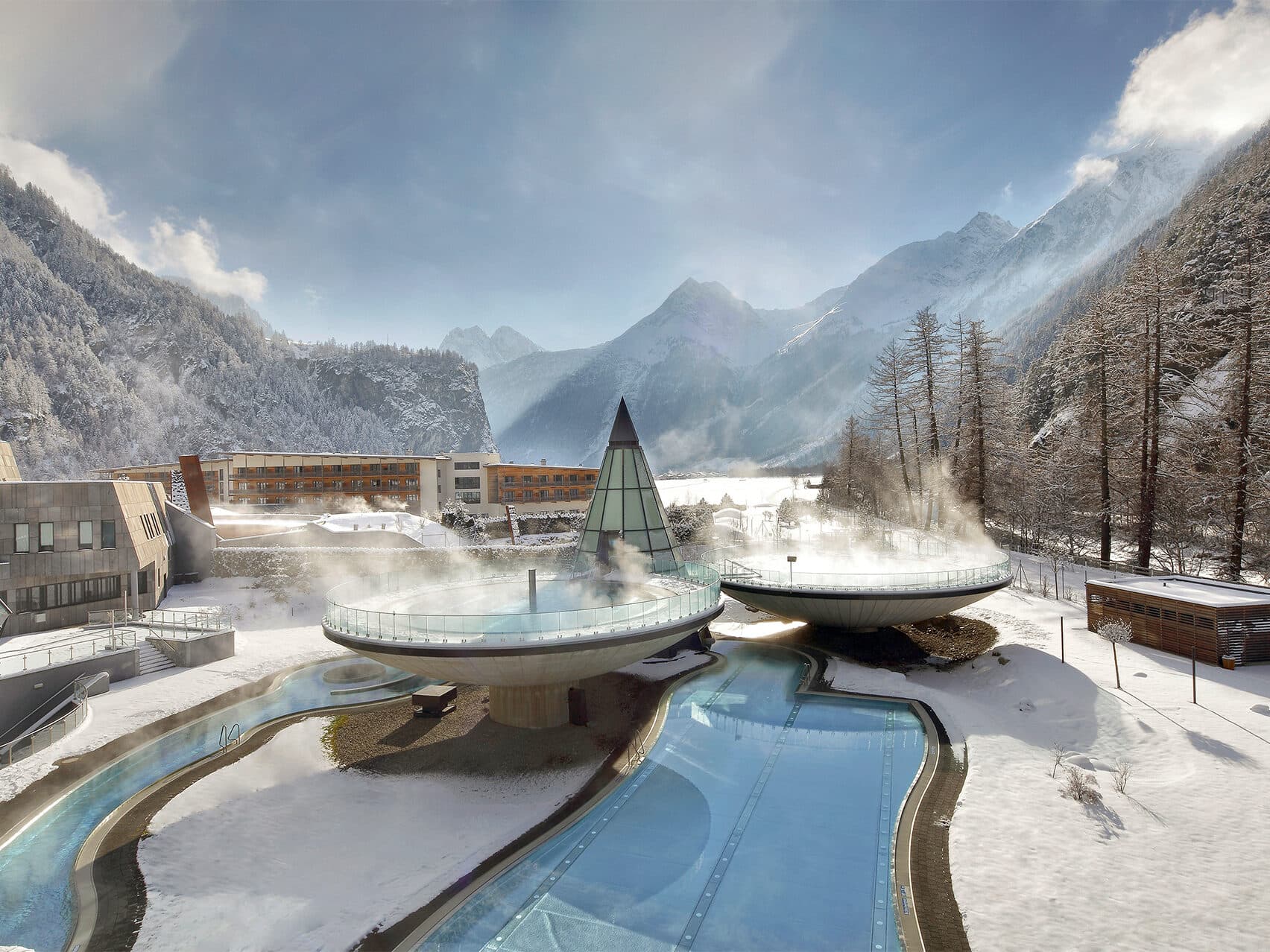 Ein modernes alpines Spa-Resort mit dampfenden Thermenbecken, eingebettet zwischen schneebedeckten Bergen und Kiefern, unter einem teilweise bewölkten Himmel.