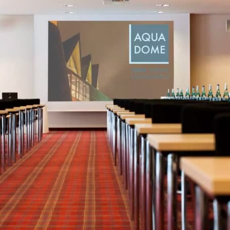 Der Konferenzraum verfügt über Tisch- und Stuhlreihen vor einem großen Bildschirm mit der Aufschrift „Aqua Dome“. Wasserflaschen sind strategisch auf den Tischen rechts platziert und schaffen so eine erfrischende Atmosphäre, die ideal ist, um geschäftliche und fitnessorientierte Gespräche zu verbinden.