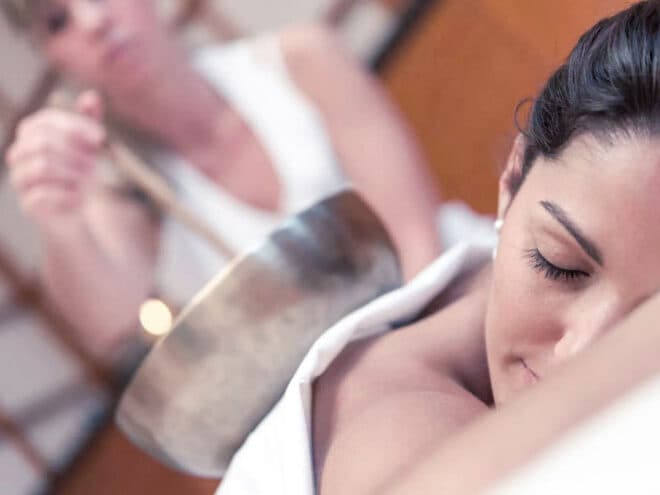 A woman relaxes at a spa, lying down with eyes closed, while another gently holds a singing bowl over her back, enveloping her in soothing sound therapy.