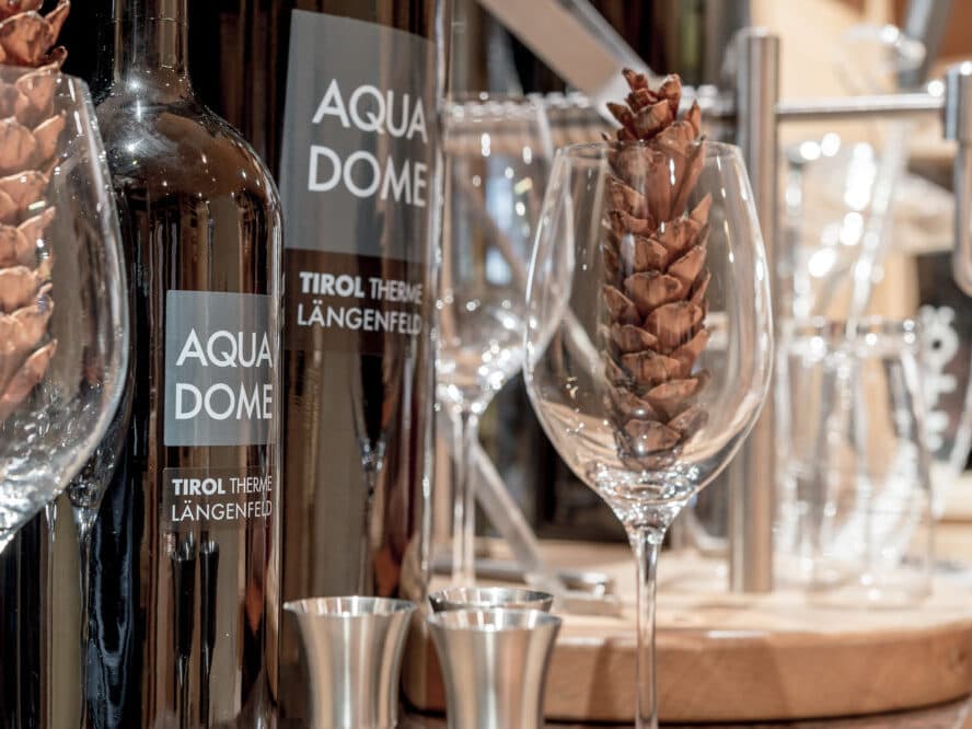 Бутылки с вином с этикеткой «Aqua Dome» и пустые бокалы с шишками внутри, выставленные на деревянной поверхности рядом с металлической посудой для питья.