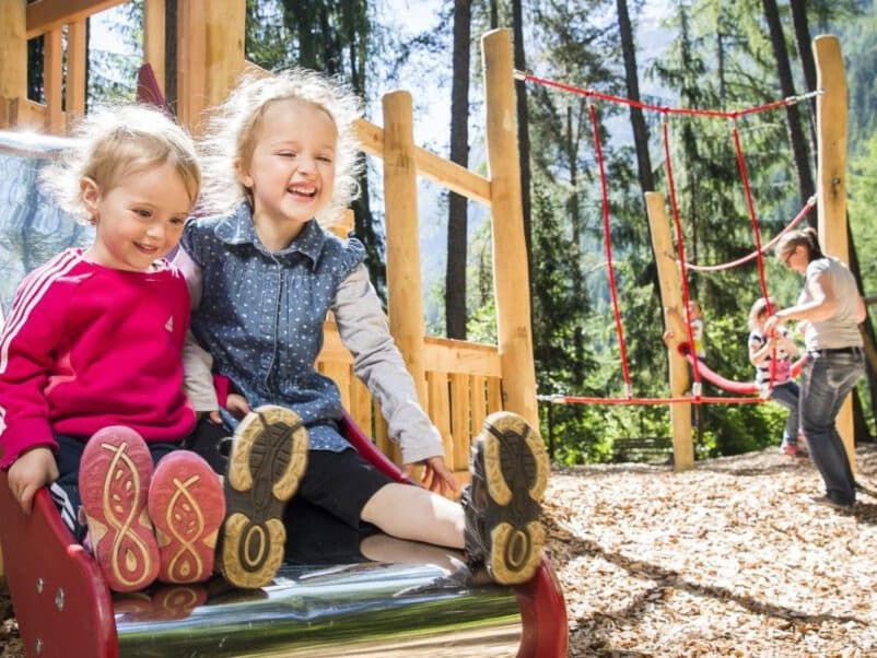 Deux jeunes filles de familles différentes dévalent un toboggan en souriant. À l'arrière-plan, un enfant grimpe à une échelle de corde sous le regard d'un adulte. De grands arbres entourent l'espace.