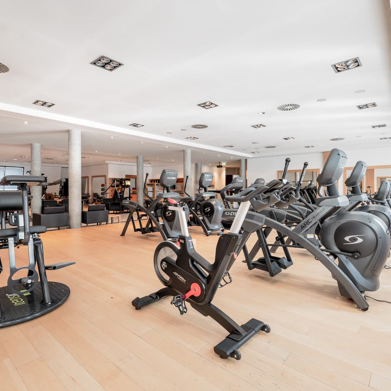 Ein geräumiges Fitnessstudio mit Holzböden, Heimtrainern, Crosstrainern und Kraftgeräten unter Deckenbeleuchtung.