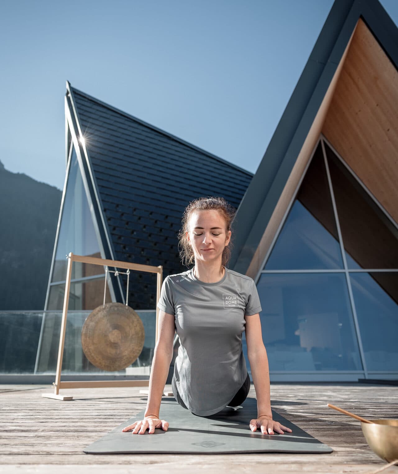 Eine Person praktiziert Yoga in der nach oben gerichteten Hundestellung auf einem Holzdeck und verkörpert damit einen ruhigen Moment der Fitness. Ein dreieckiges Dach und ein Gong tragen im Hintergrund zur ruhigen Atmosphäre bei.