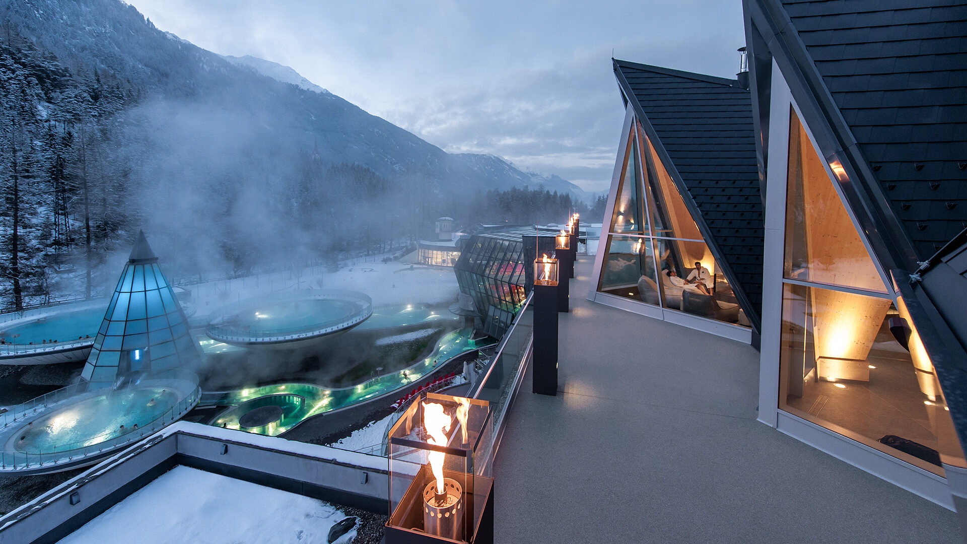 Mountain resort view with modern architecture, featuring illuminated triangular glass structures, steaming spa pools, and snow-covered peaks. Fire pits gracefully line the balcony railing in the foreground.