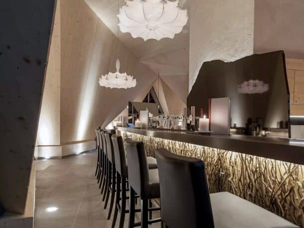 The modern bar interior, featuring high-backed chairs along a sleek counter, offers a spa-like tranquility. Unique geometric ceiling designs paired with artistic light fixtures complete the serene ambiance.