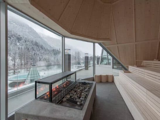 Modern indoor sauna with large windows overlooking a snowy landscape, creating a spa-like retreat. Featuring wooden benches and a rectangular central fire pit.