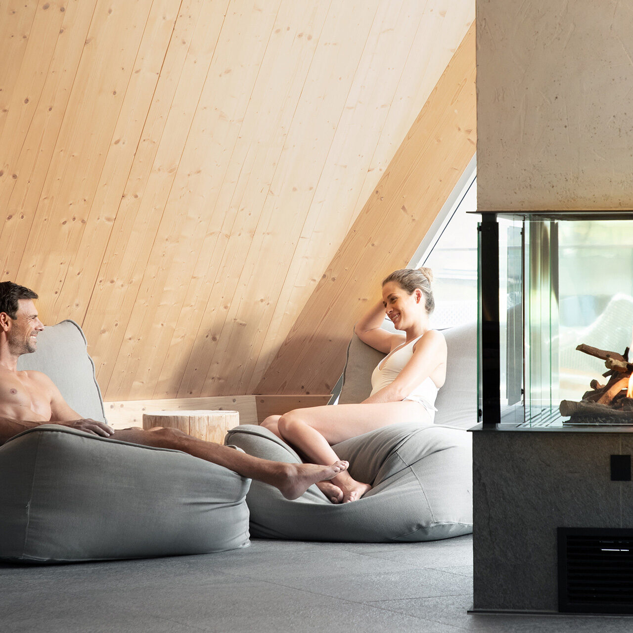 Zwei Personen entspannen auf grauen Sitzsäcken in einem gemütlichen Raum mit Holzwänden und einem verglasten Kamin.